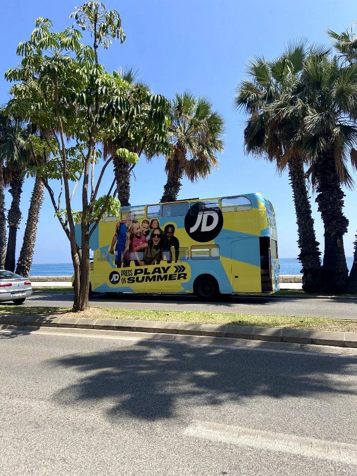 El autobús de dos plantas de JD, rotulado con Press Play on Summer, recorriendo Málaga junto al mar.