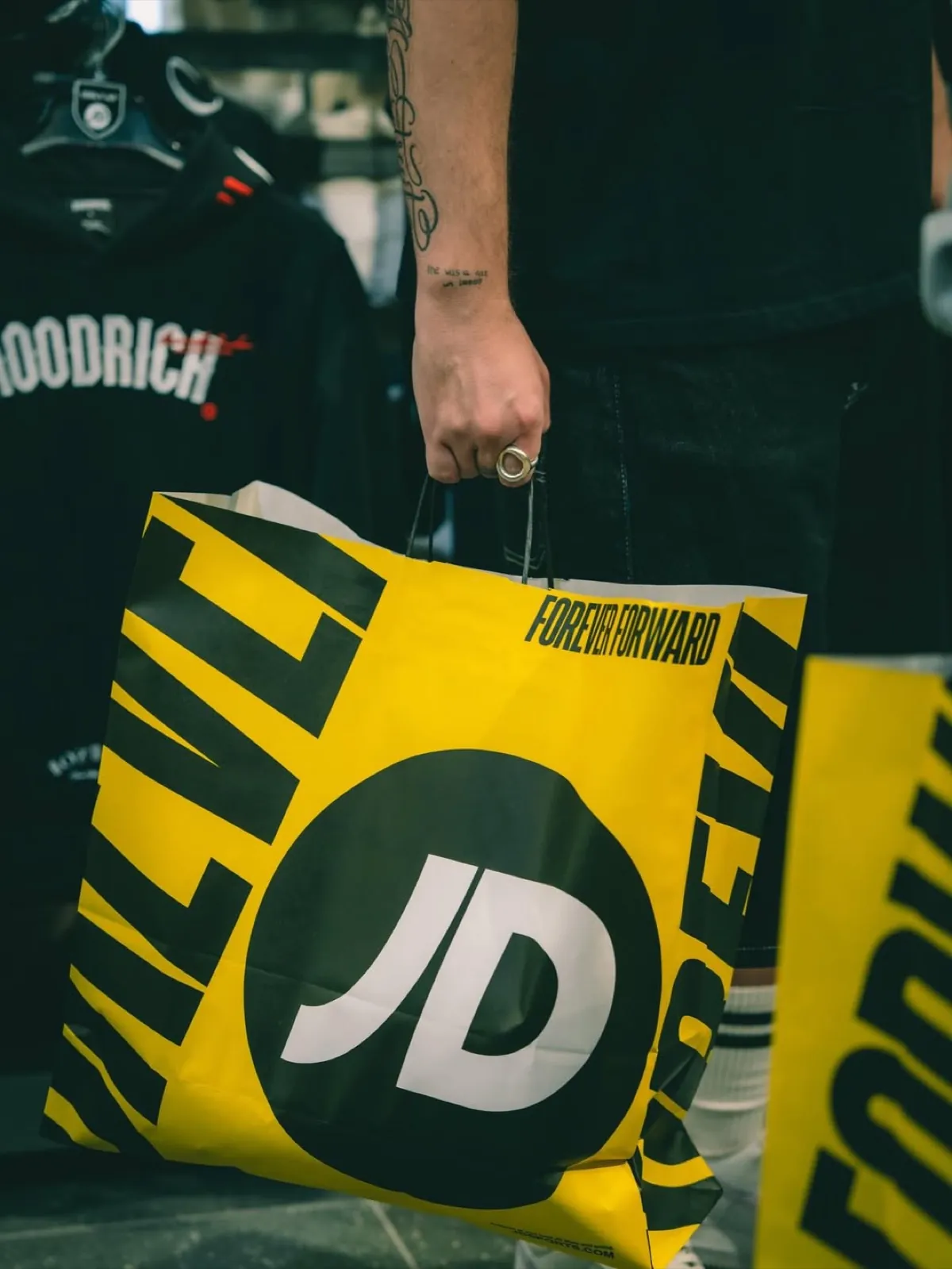 Bolsa de JD con el lema «Forever Forward» en la tienda, junto a ropa de la marca.