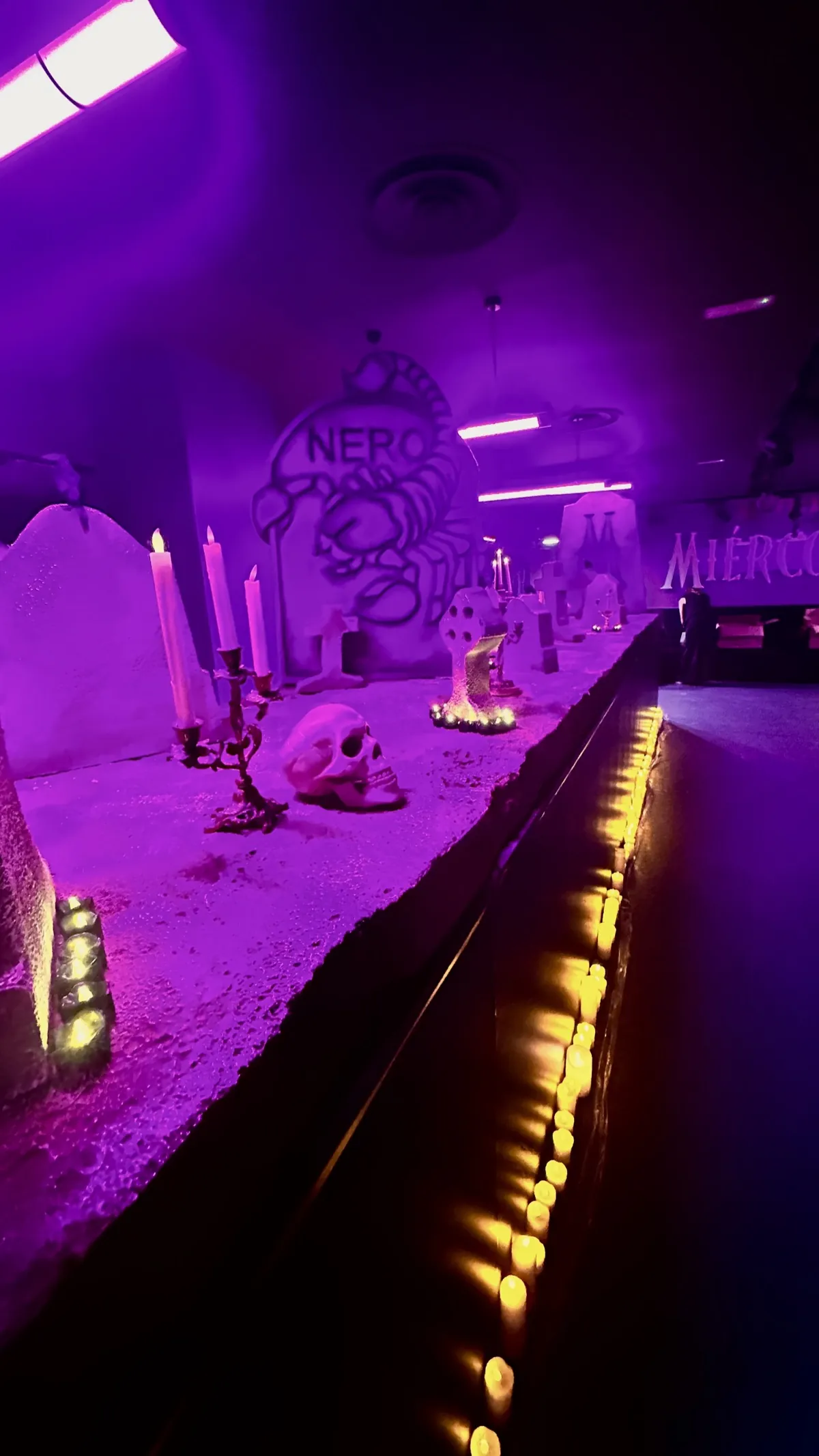 Interior del restaurante transformado en un cementerio: lápidas, candelabros, velas y los rótulos de Miércoles iluminados en púrpura.