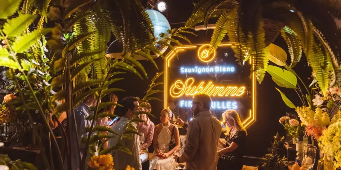 Rótulo luminoso de Sublimme entre plantas de la floristería, con invitados alrededor.